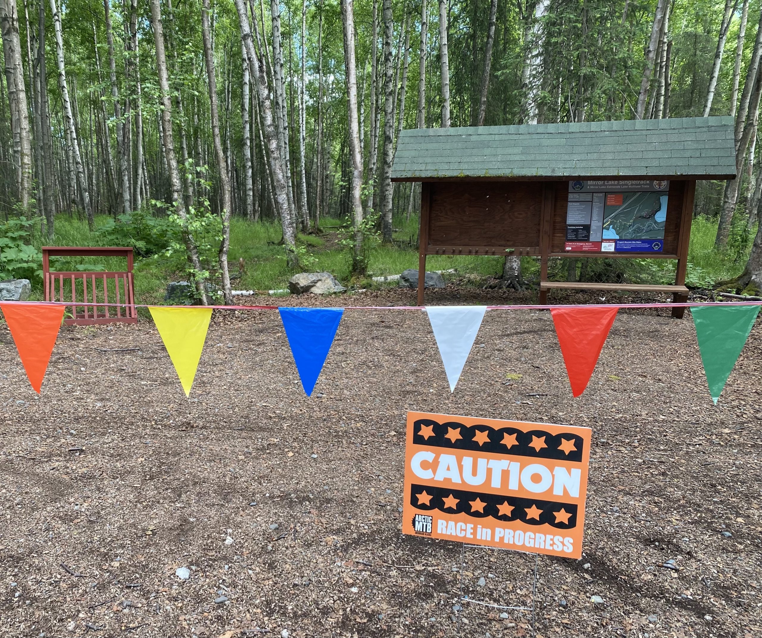 A forested area with a wooden information kiosk in the background, partially hidden by trees. In the foreground, a brightly colored banner of triangular flags in orange, yellow, blue, and white stretches across the scene. A sign reads "CAUTION RACE in PROGRESS," indicating that an event is taking place. The ground is covered in dirt and small rocks, with scattered greenery around. Mirror Lake mountain bike trail.