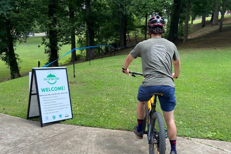 A person wearing a helmet is on a yellow mountain bike, facing a welcome sign at a pop-up bike park. The sign outlines trail rules and nearby greenery, with a pathway leading downhill.