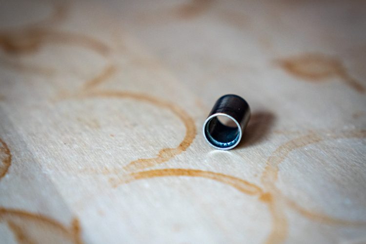 A close-up view of a small metallic tube lying on a wooden surface marked with circular stains. The tube is cylindrical, with a shiny silver rim and a darker center.