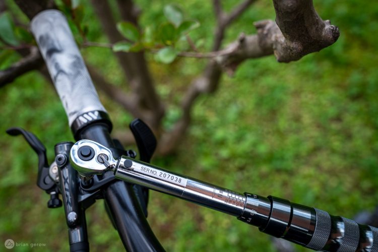 Close-up view of a bicycle handlebar featuring a ratchet tool and a brake lever. The handlebar has a textured grip, and the background is a blurred green landscape with tree branches. The ratchet tool has a visible serial number and measurement markings.