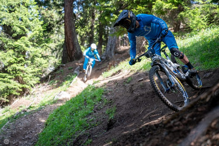 Two mountain bikers in blue attire navigate a dirt trail surrounded by trees. The foreground rider leans into a turn, while the second rider is farther down the path. Sunlight filters through the trees, highlighting the lush green grass and dirt trail.