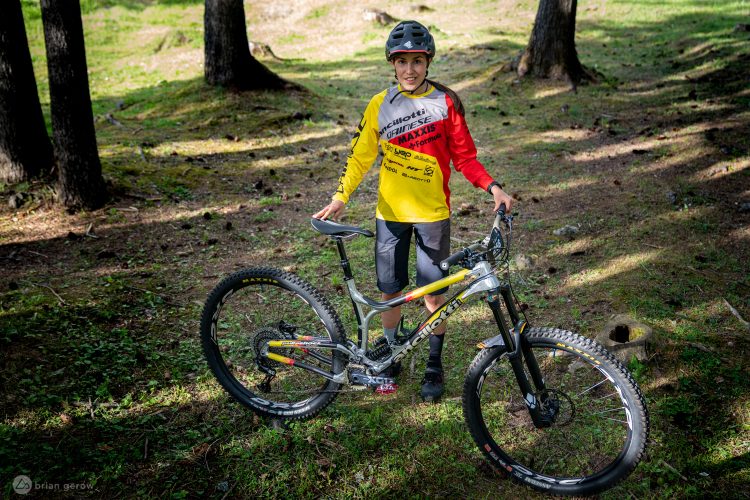 A young cyclist in a bright red and yellow jersey stands next to a mountain bike in a forested area. The cyclist is wearing a helmet and shorts, with trees and grass visible in the background. The scene captures a moment of excitement and readiness for biking.