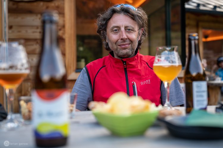 A man in a red jacket sits at a table with various beverages, including a glass of beer and bottles. In front of him are snacks, including chips, with a rustic wooden background in a casual outdoor setting.
