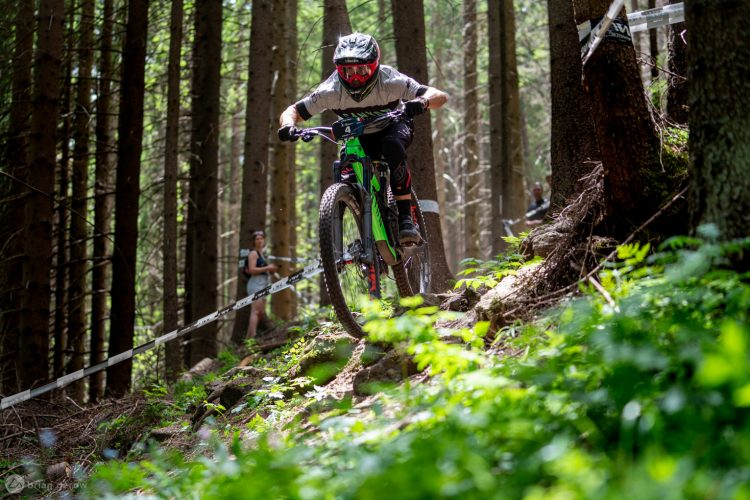 A mountain biker in a helmet and protective gear descends a rocky trail in a dense forest, surrounded by tall trees and greenery. In the background, a spectator watches the race, capturing the excitement of the outdoor event.