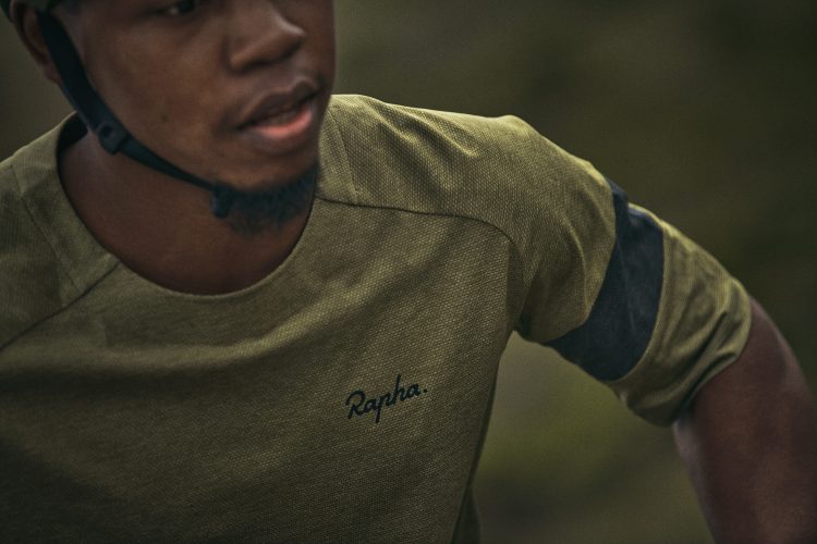 A close-up image of a man wearing a green athletic shirt with the brand name "Rapha" embroidered on it. He is partially turned, wearing a black helmet, with a blurred outdoor background.
