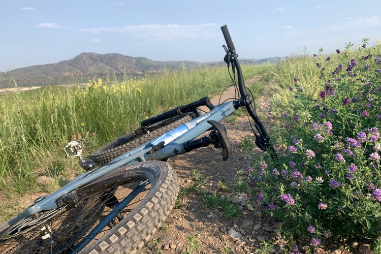 A mountain bike lies tilted on a dirt path surrounded by tall grass and blooming purple flowers, with rolling hills in the background under a clear blue sky.