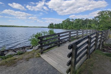 A wooden bridge extends over calm water, surrounded by lush greenery and a cloudy blue sky. The scene captures a peaceful lakeside view with rocks lining the shore and trees in the background. Manasquan Reservoir mountain bike trail.