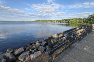 A scenic view of a calm lake surrounded by lush green trees under a partly cloudy sky. In the foreground, a wooden walkway leads along the water’s edge, with large, smooth stones along the shore. The reflection of the trees and clouds can be seen in the still water, creating a peaceful atmosphere. Manasquan Reservoir mountain bike trail.