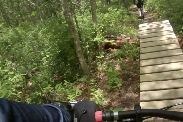 A close-up view of a mountain biker's handlebars and arm, riding on a wooden bridge through a lush, green forest. A second biker is visible in the background, following the trail. Seaview Trails Main loop mountain bike trail.
