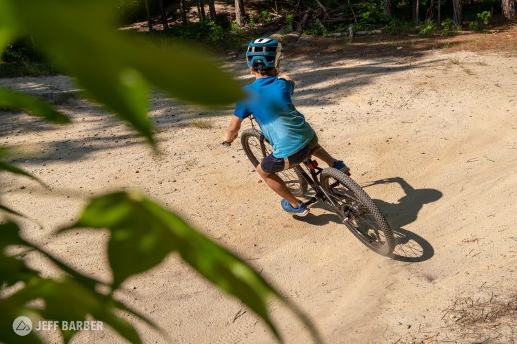 A child riding a mountain bike on a sandy trail, surrounded by trees, with sunlight filtering through the leaves. The child is wearing a blue helmet and a blue shirt, showcasing a sense of adventure and outdoor activity.