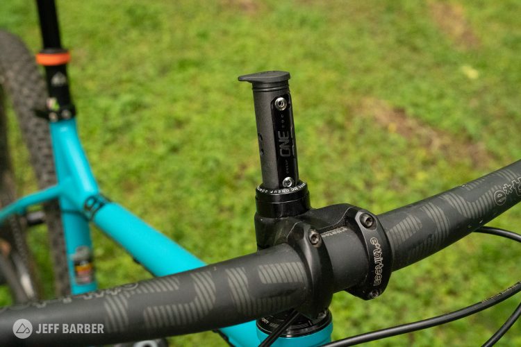 Close-up view of a mountain bike's handlebar setup, featuring a black stem with a digital display mount, a turquoise frame, and a dropper post. The background shows a grassy area, emphasizing an outdoor cycling environment.
