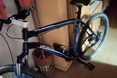 Specialized Rockhopper Comp Disc: A blue and black mountain bike is positioned in a room, with various household items visible in the background. The bike features front suspension and knobby tires, indicating it is designed for off-road riding. The room has warm-colored walls and some clothing and storage furniture is seen in the surroundings.