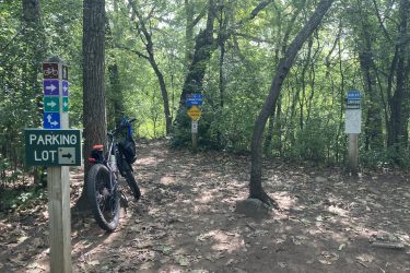 A dirt path in a wooded area, featuring several directional signs for trails, a "Parking Lot" sign pointing to the right, and a mountain bike leaned against a tree. The scene is surrounded by lush greenery and fallen leaves on the ground. Kettle Moraine John Muir + Emma Carlin mountain bike trail.