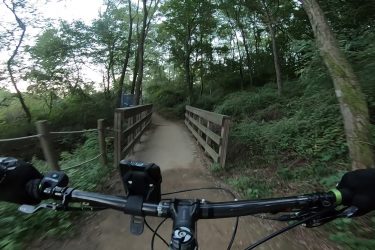A view from the handlebars of a mountain bike, showing a dirt path leading to a wooden bridge surrounded by lush greenery and trees, indicating a trail in a natural setting. Gyeyangsan mountain bike trail.