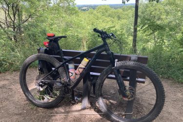 A mountain bike is positioned next to a wooden bench, set against a backdrop of lush greenery and rolling hills in the distance. The bike features thick tires and a water bottle attached, indicating it is ready for an outdoor adventure. Kettle Moraine John Muir + Emma Carlin mountain bike trail.