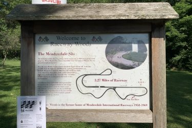 A wooden sign at Raceway Woods welcomes visitors and provides historical information about the site, including details about the Meadowdale Silo and the former Meadowdale International Raceway, which operated from 1958 to 1969. The sign features a map of the 3.27-mile racetrack and highlights the area's natural beauty. Alongside the sign is a flyer for a 5K challenge event taking place in August 2023. Background features lush greenery typical of a wooded area. Raceway Woods mountain bike trail.