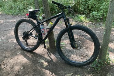 A black mountain bike parked beside a tree on a dirt trail, surrounded by lush greenery. The bike features thick tires and has two water bottles attached to the frame. Sunlight filters through the trees, creating a natural outdoor setting. John Muir Trails mountain bike trail.