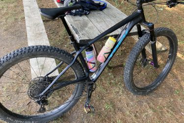 A black mountain bike is parked next to a wooden picnic table. The bike features thick, knobby tires, a sturdy frame, and a colorful water bottle attached to the frame. In the background, there is a patch of grass with some dry leaves, and a small pile of a rider's gear can be seen on the table. John Muir Trails mountain bike trail.