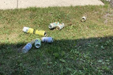 A scattered assortment of empty beverage cans resting on a patch of grass, with some partially shaded by a nearby structure. Alpine Valley mountain bike trail.