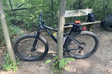 A black mountain bike with thick tires is parked next to a wooden bench in a wooded area. The bench features the engraving "Lake Country Rotary." Nearby, there are patches of greenery and a tire resting on the ground. The scene conveys a tranquil outdoor setting for cycling and relaxation. Kettle Moraine John Muir + Emma Carlin mountain bike trail.