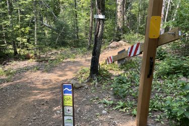Alt text: A forested trail entrance marked by a wooden post indicating directions and trail usage. Signs on the post suggest a biking trail named "Jackpot." The trail is surrounded by lush greenery and a rustic dirt path leading into the woods. A barrier with red and white stripes is partially visible along the path. Jackpot mountain bike trail.