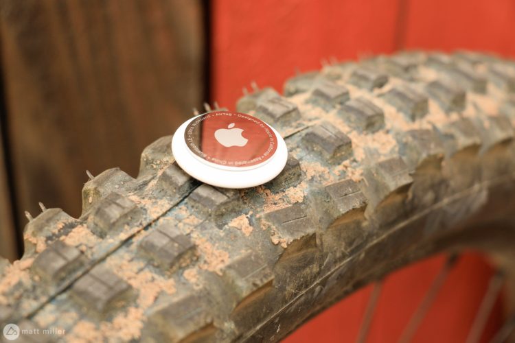 A close-up image of a mountain bike tire with dirt and grime on it, featuring an Apple AirTag resting on the tire tread. The background includes a wooden surface painted in a shade of red.