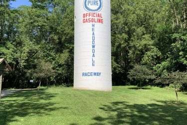 A tall, cylindrical gas storage tank with a white exterior, featuring the blue and red text "PURE OFFICIAL GASOLINE MEADOWDALE RACEWAY." The tank is surrounded by lush green grass and trees under a clear blue sky. Raceway Woods mountain bike trail.