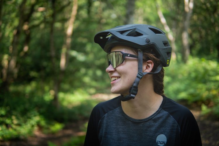 A smiling individual wearing a gray biking helmet and sunglasses, standing in a lush green forest setting. The person is dressed in a dark athletic shirt, highlighting a sense of outdoor adventure.
