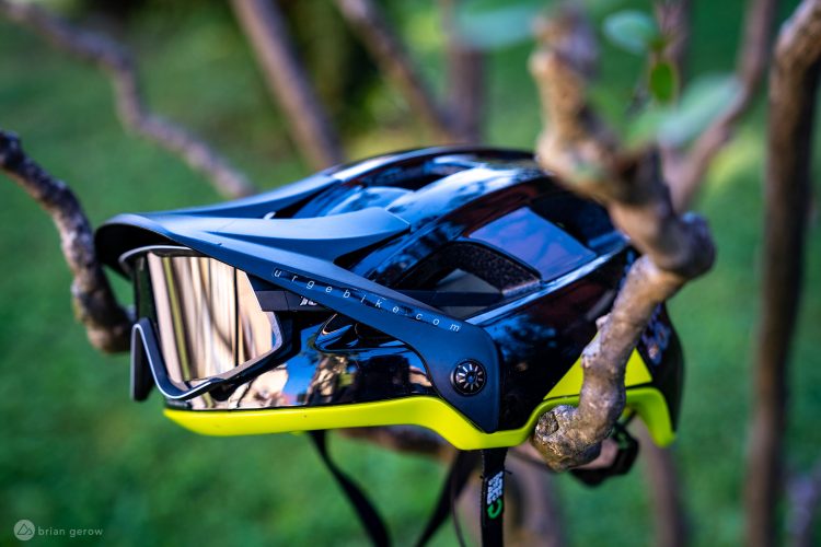 A black and yellow mountain biking helmet hanging on a tree branch, featuring a visor and reflective sunglasses attached, with greenery in the background.