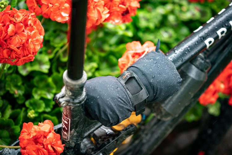 A close-up of a bike handlebar covered with a waterproof cover, resting near vibrant orange flowers. Rain droplets can be seen on the cover and surrounding foliage, indicating a wet environment.