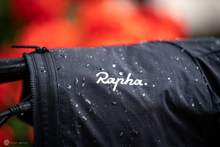 Close-up of a black cycling jacket with the brand name "Rapha" prominently displayed. The fabric is showing droplets of water, indicating its water-resistant quality, with a blurred background featuring red and green colors.