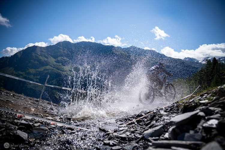 A mountain biker splashes through a stream of water on a rocky trail, with majestic mountains and a blue sky in the background. Sunlight glints off the water, creating a dynamic and adventurous scene.