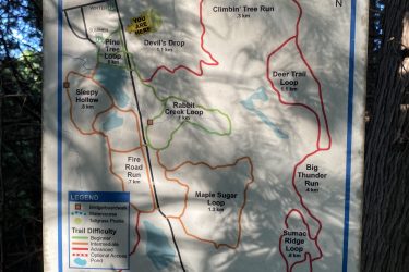 Map of the Allan Park Trails, displaying various hiking routes and their distances, including labels for trail names like "Devil's Drop," "Maple Sugar Loop," and "Big Thunder Run." The map indicates trail difficulty levels and features such as bridges and watercourses, along with a "You Are Here" marker for orientation. The background consists of trees, suggesting a natural outdoor setting. Allen Park mountain bike trail.