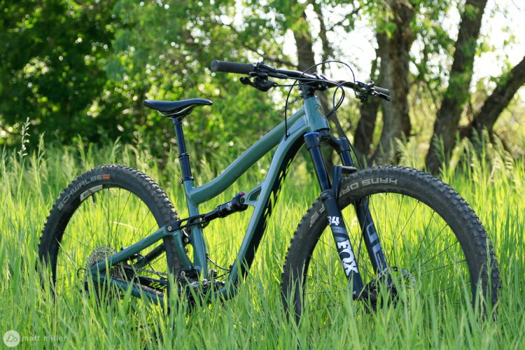 A mountain bike leaning against tall green grass, surrounded by trees in a natural outdoor setting. The bike features a sleek frame and prominent tires designed for trail riding.