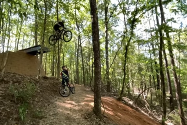 A mountain biker performs a jump above the trail in a wooded area, while another biker looks on. The scene is filled with green trees and sunlight filtering through the leaves, highlighting the action and excitement of mountain biking in nature. Bogue Chitto State Park mountain bike trail.
