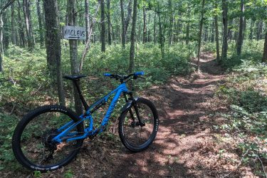 A blue mountain bike is parked near a sign labeled "NUCLEUS" pointing toward a wooded trail. The scene features a lush green forest with tall trees, undergrowth, and a winding dirt path, indicating a tranquil outdoor setting ideal for biking or hiking. Seaview Trails Main loop mountain bike trail.