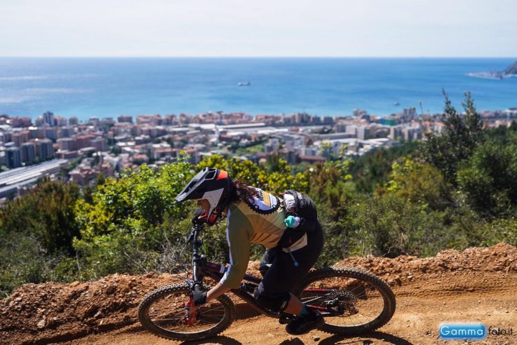 A mountain biker navigating a dirt trail with a scenic view of a coastal city and the ocean in the background. The cyclist is wearing protective gear, including a helmet, and appears focused as they ride downhill. Lush greenery lines the path, contrasting with the urban landscape below.