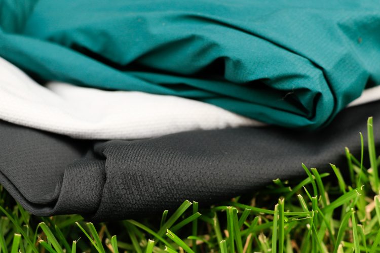 Three folded pieces of fabric in varying colors—teal, white, and black—rest on a bed of green grass. The textures of the fabrics are visible, showcasing their smooth surfaces.