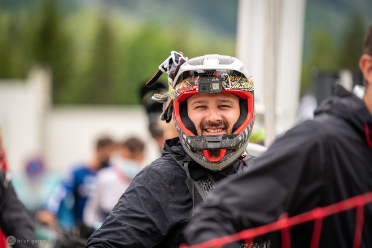 A smiling man in a protective mountain biking helmet and gear, standing in a crowd at an outdoor event, with trees and blurred spectators in the background.