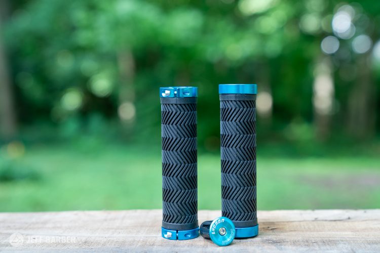 Two bicycle handlebar grips with textured black rubber and vibrant blue aluminum end caps, placed on a wooden surface against a blurred green background.