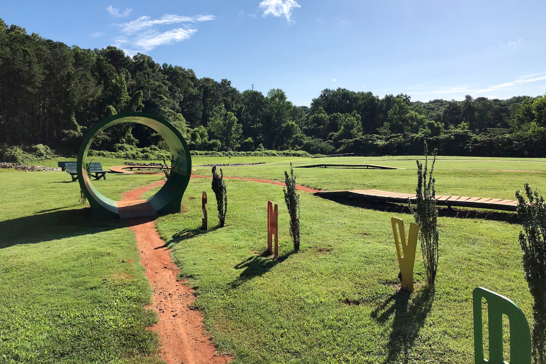 A serene park scene featuring a winding dirt path leading through a grassy area. A large green circular archway is visible in the foreground, inviting visitors to walk through. Colorful sculptures in the shape of letters are scattered throughout the area, which is framed by lush trees in the background and a bright blue sky overhead. Benches can be seen off to the left, providing spots for relaxation. North Cooper Lake Park Trail mountain bike trail.