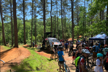 A lively outdoor scene featuring a group of cyclists and spectators gathered in a wooded area. The background includes tall trees and dirt bike trails, while a van with an open back is visible, likely providing refreshments or support. Various individuals, including children and adults, are interacting, some wearing helmets and cycling gear in preparation for biking activities. Bright sunlight filters through the trees, creating a vibrant atmosphere. Bogue Chitto State Park mountain bike trail.