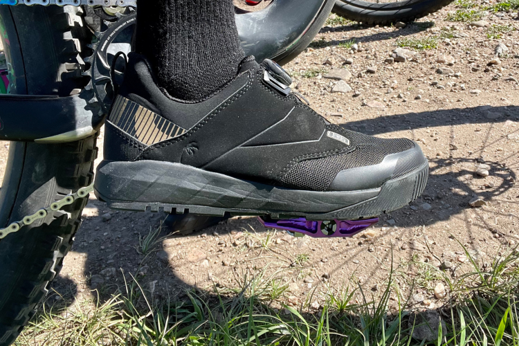 A close-up view of a black cycling shoe attached to a purple pedal, with a bicycle chain visible in the background. The shoe features a textured design and a snug fit, resting on a dirt surface with some grass.
