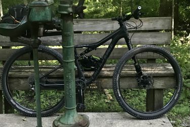 A mountain bike parked next to an old, green hand pump on a concrete platform surrounded by trees and vegetation. A wooden bench is in the background, adding to the rustic outdoor setting. Greenbrier River Trail mountain bike trail.