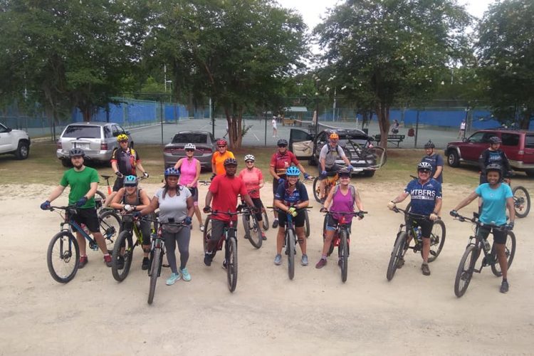 A diverse group of individuals, both male and female, are gathered on bicycles in a park setting. They are wearing helmets and athletic clothing, ready for an outdoor cycling activity. In the background, there are trees and parked vehicles, along with tennis courts visible in the distance. The scene conveys a sense of community and enthusiasm for biking.