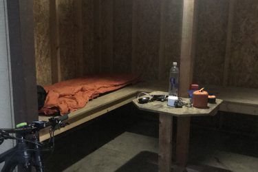 Interior of a small shelter featuring a wooden bench with an orange sleeping bag, a wooden table with various items, and a bicycle parked in the corner. The space is illuminated by a light on the wall, with walls of plywood and a simple wooden floor. Greenbrier River Trail mountain bike trail.