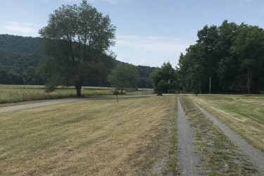 A scenic view of a grassy field bordered by trees and rolling hills under a partly cloudy sky. A gravel path runs through the center, leading towards the horizon. Greenbrier River Trail mountain bike trail.