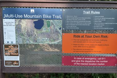 Signboard displaying information about the Multi-Use Mountain Bike Trail at Lone Lake Park. The sign includes a map of the trail, hours of operation, and important trail rules such as controlling bicycles, respecting other users, and riding at your own risk. Additional warnings about hazards and contact information for emergencies are also provided. Lone Lake Park mountain bike trail.