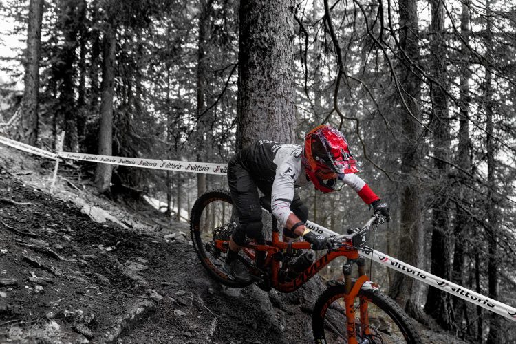 A mountain biker navigating a rocky downhill trail in a forested area, with a focus on the rider in vibrant red gear and an orange bike against a monochromatic background of trees and dirt. A safety tape is visible in the scene, indicating a racing or practice course.