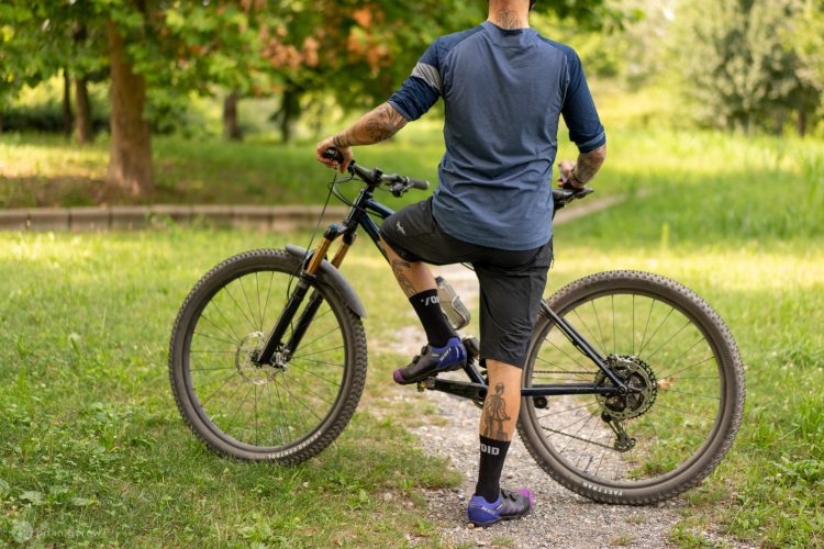 A person standing next to a mountain bike on a gravel pathway in a park. The individual is wearing a blue long-sleeve shirt and black shorts, with colorful cycling shoes and knee-high socks. They have tattoos visible on their arms and legs. The background features green trees and grass, indicating a sunny day outdoors.